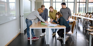 Studierende Gruppenarbeit Studierende stehen gemeinsam um einen Tisch im Seminarraum und arbeiten.