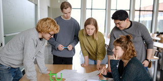 Studierende Gruppenarbeit Fünf Studierende stehen um einen Tisch und sehen auf ein Dokument.